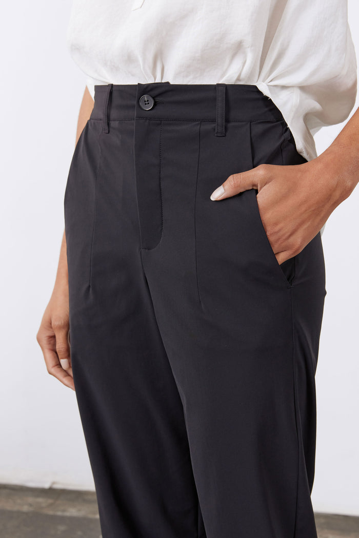 Black tapered pants close-up: button waistband, pleats, and front seam.